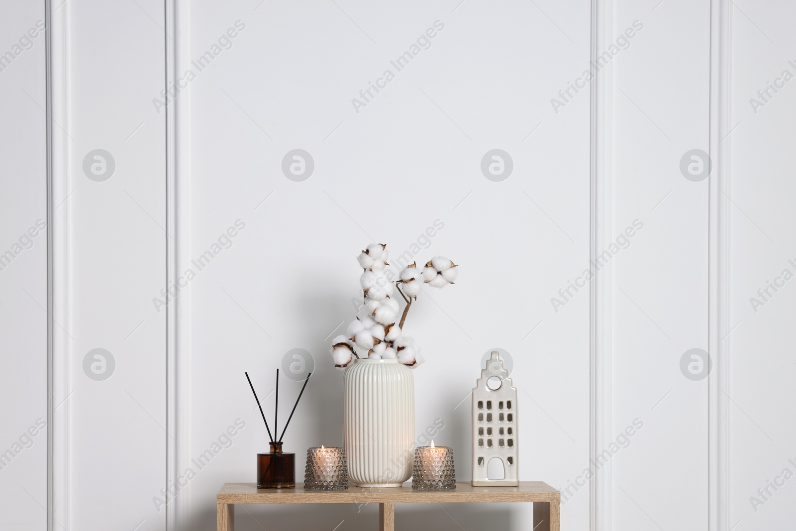 Candlesticks with burning candles and other decor on wooden table near white wall indoors, space for text Photo of Candlesticks with burning candles and other decor on wooden table near white wall indoors, space for text