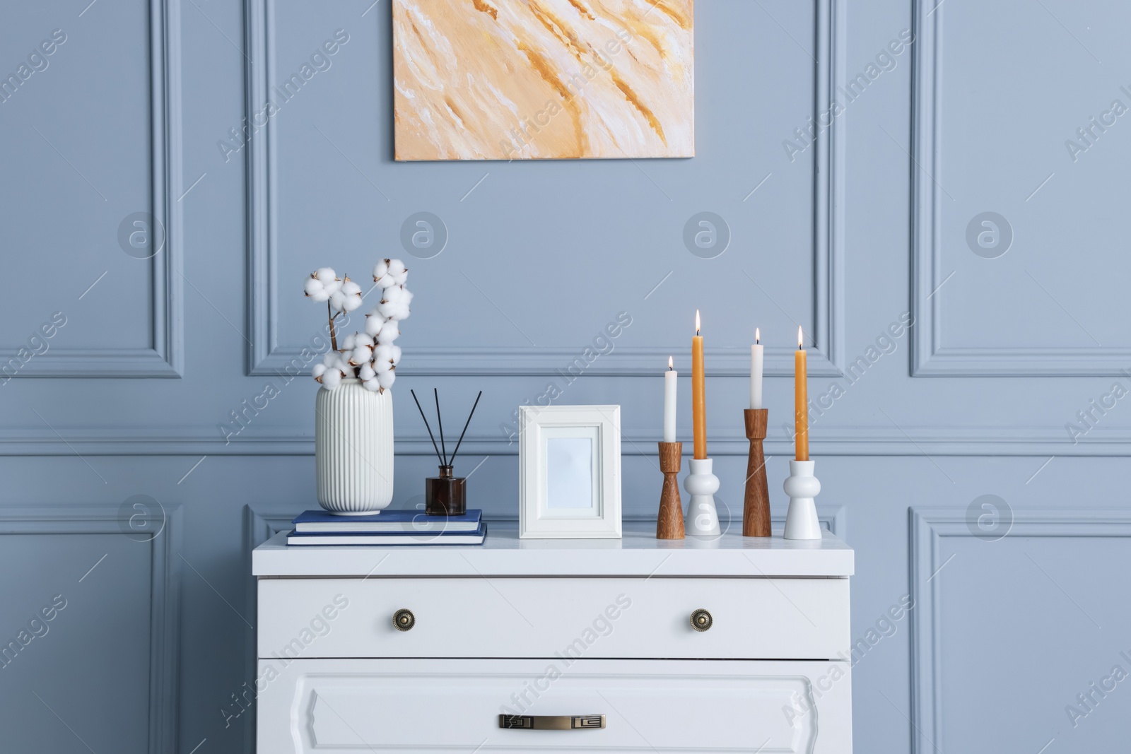 Stylish candlesticks with burning candles, cotton flowers in vase and other decor on white chest of drawers near gray wall indoors Photo of Stylish candlesticks with burning candles, cotton flowers in vase and other decor on white chest of drawers near gray wall indoors