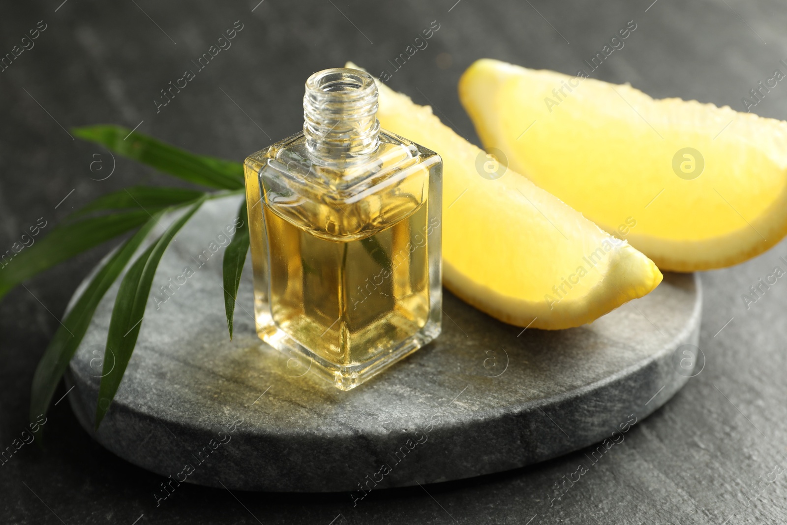 Photo of Open perfume bottle with aromatic essential oil, leaves and slices of lemon on dark textured table, closeup