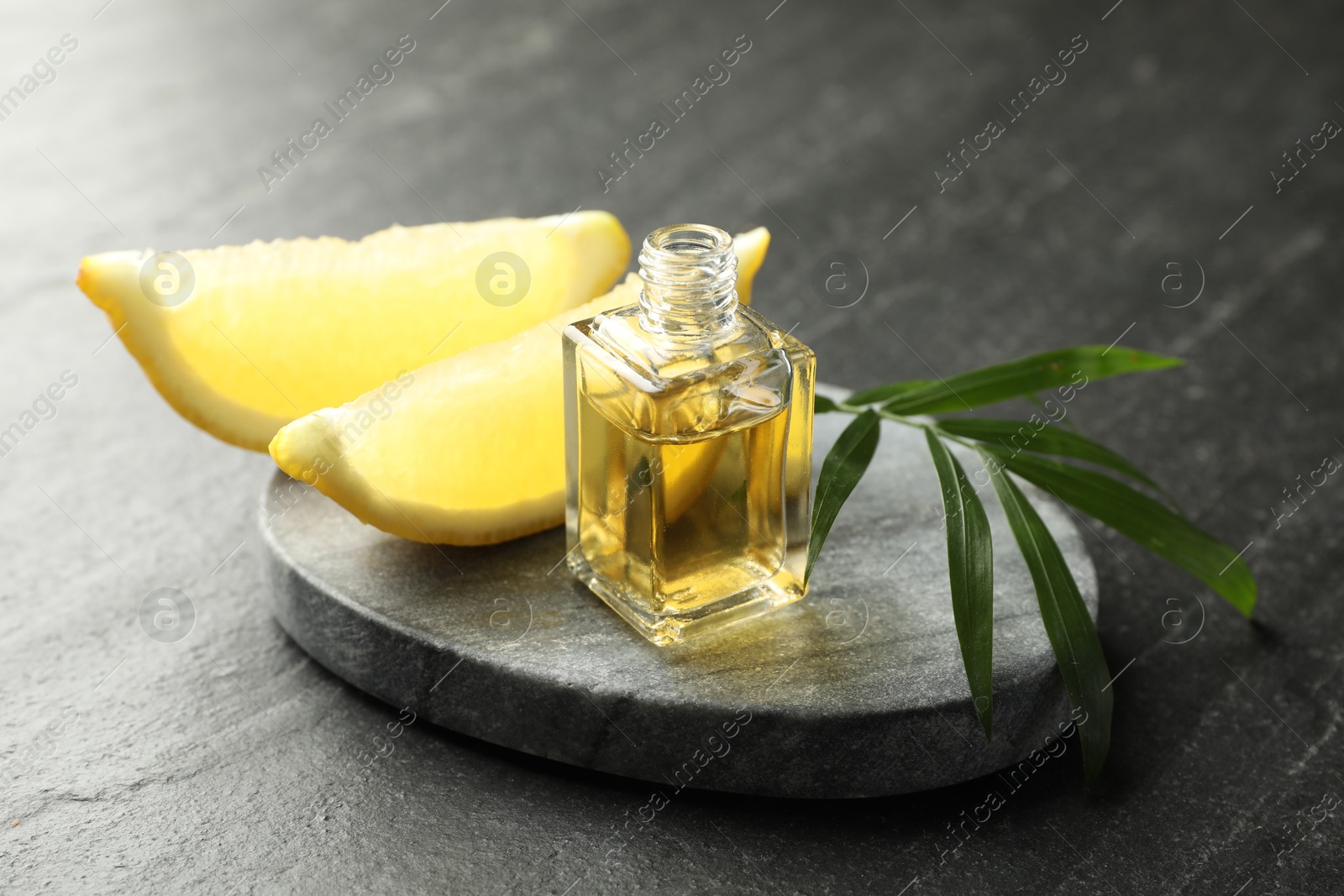 Photo of Open perfume bottle with aromatic essential oil, leaves and slices of lemon on dark textured table, closeup