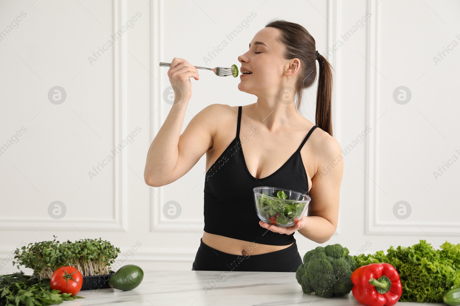 Woman in fitness clothes eating salad at white marble table indoors. Healthy food Photo of Woman in fitness clothes eating salad at white marble table indoors. Healthy food
