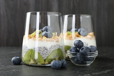 Photo of Delicious dessert with chia seeds, yogurt, granola, blueberries and kiwi in glasses on black table, closeup. Superfood