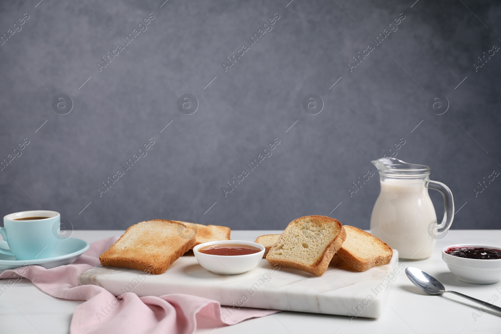 Slices of tasty toasted bread, milk, coffee and jam on white table against grey background. Space for text Photo of Slices of tasty toasted bread, milk, coffee and jam on white table against grey background. Space for text