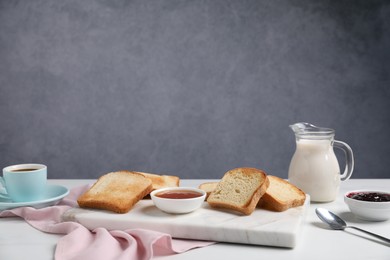 Photo of Slices of tasty toasted bread, milk, coffee and jam on white table against grey background. Space for text