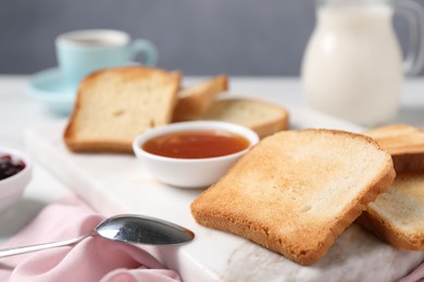 Photo of Slices of tasty toasted bread, spoon and jam on white table against grey background, closeup