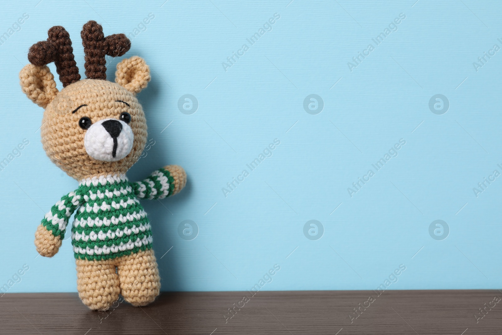 Photo of Cute knitted toy deer on wooden table against light blue background. Space for text