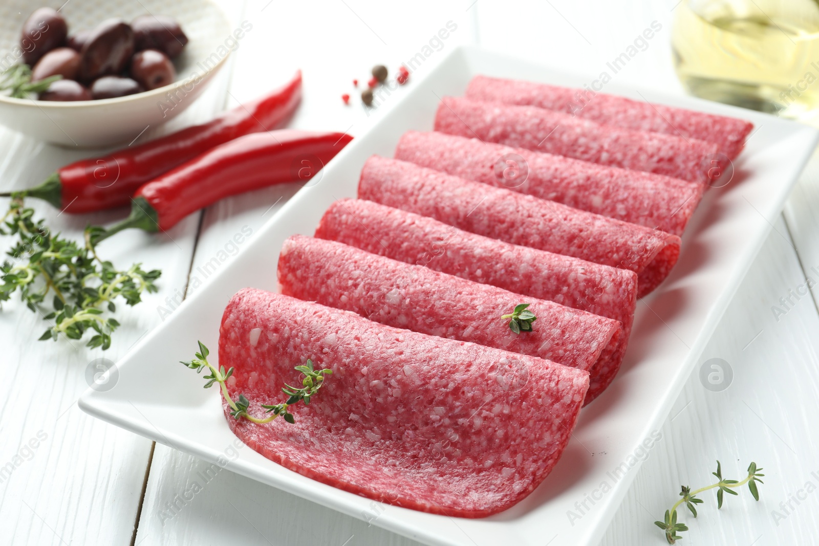 Photo of Slices of tasty smoked sausage, spices and olives on white wooden table, closeup