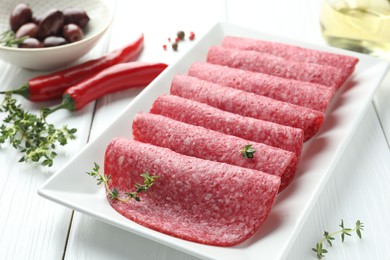 Photo of Slices of tasty smoked sausage, spices and olives on white wooden table, closeup