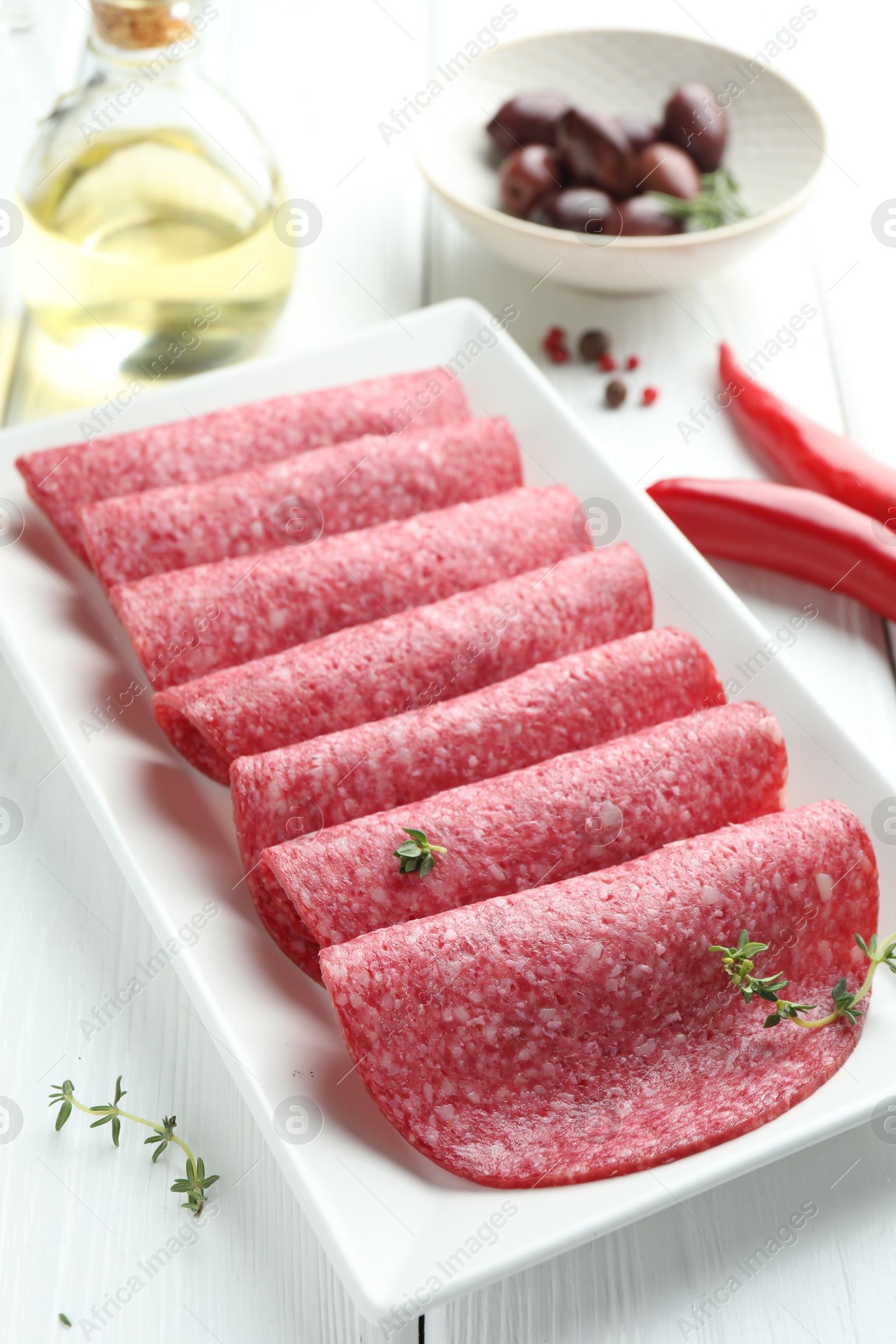 Photo of Slices of tasty smoked sausage, spices and olives on white wooden table, closeup