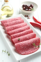 Photo of Slices of tasty smoked sausage, spices and olives on white wooden table, closeup