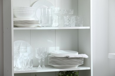 Photo of Clean glasses and plates on shelves near white wall indoors