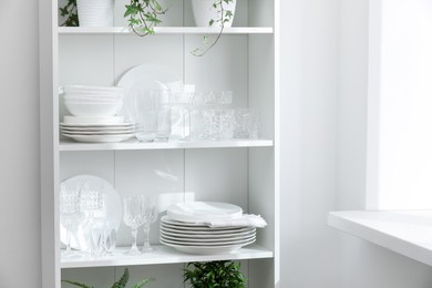 Photo of Clean glasses, plates and green houseplants on shelves near white wall indoors
