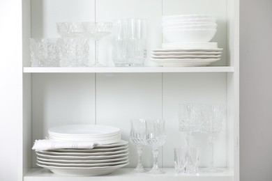 Photo of Clean glasses and plates on shelves near white wall indoors