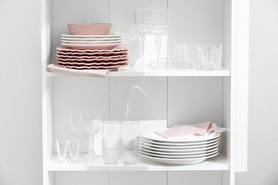 Photo of Clean glasses and plates on shelves near white wall indoors