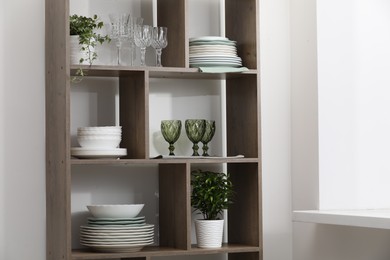 Photo of Clean glasses, plates and green houseplants on shelves near white wall indoors