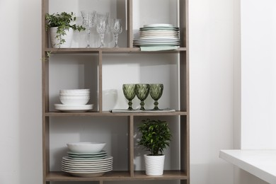 Photo of Clean glasses, plates and green houseplants on shelves near white wall indoors