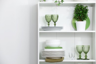 Photo of Clean glasses, plates and green houseplant on shelves near white wall indoors, space for text