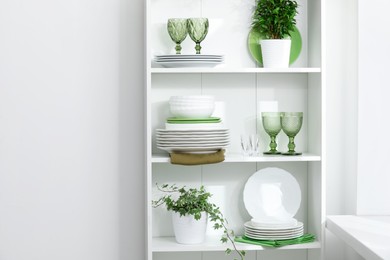 Photo of Clean glasses, plates and green houseplants on shelves near white wall indoors, space for text
