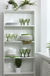 Photo of Clean glasses, plates and green houseplants on shelves near white wall indoors