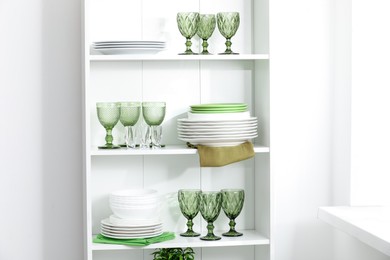 Photo of Clean glasses and plates on shelves near white wall indoors