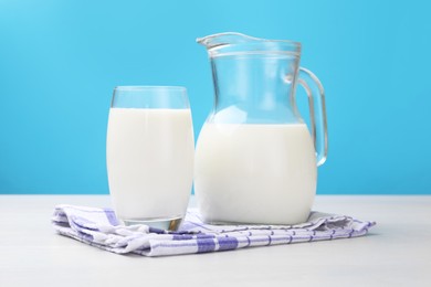 Photo of Fresh milk in glass and jug on white wooden table against light blue background