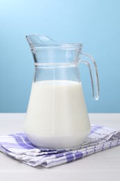 Photo of Fresh milk in jug on white wooden table against light blue background