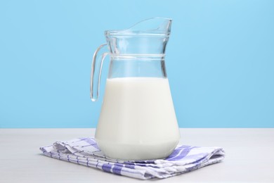 Photo of Fresh milk in jug on white wooden table against light blue background