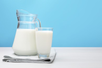 Photo of Fresh milk in glass and jug on white wooden table against light blue background. Space for text
