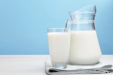 Photo of Fresh milk in glass and jug on white wooden table against light blue background. Space for text