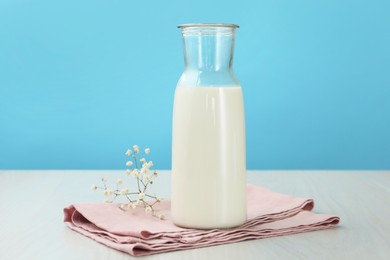 Photo of Fresh milk in jug and flowers on white wooden table against light blue background