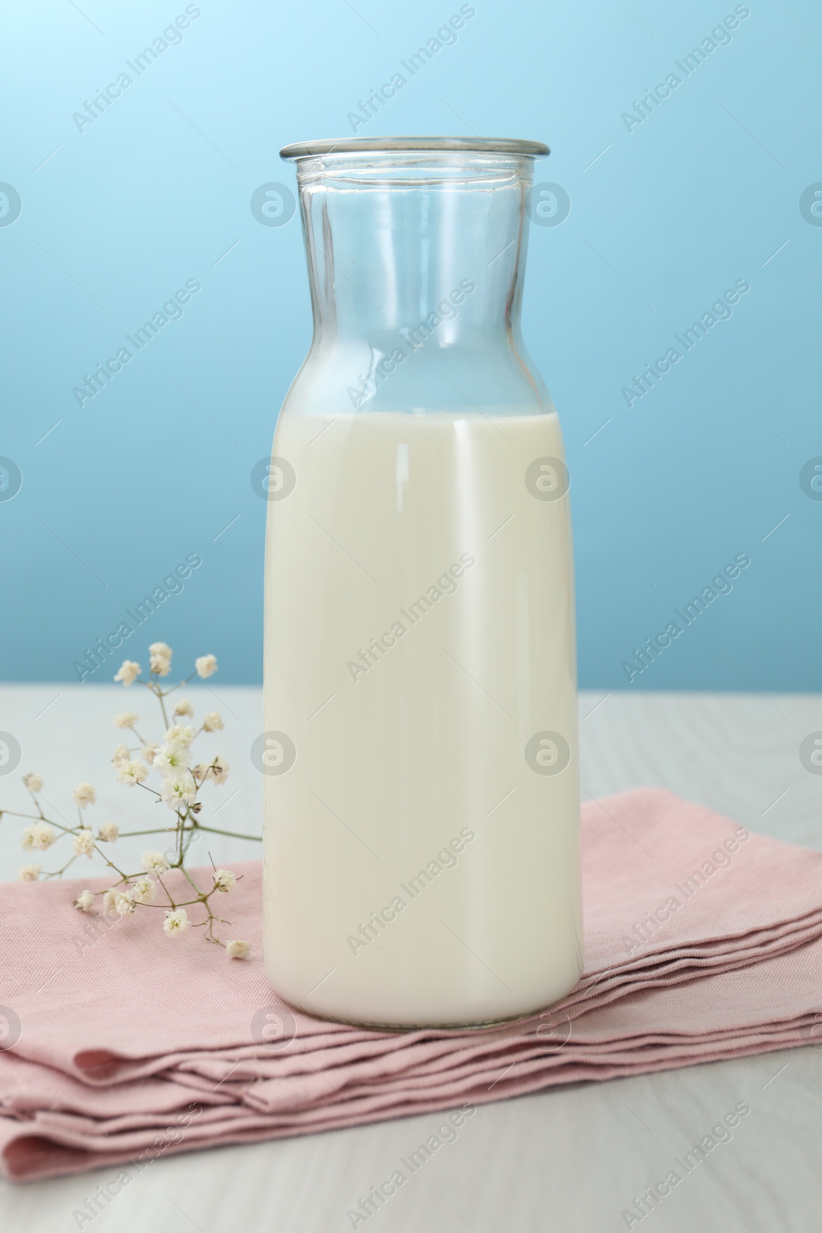 Fresh milk in jug and flowers on white wooden table against light blue background Photo of Fresh milk in jug and flowers on white wooden table against light blue background