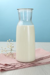 Photo of Fresh milk in jug and flowers on white wooden table against light blue background