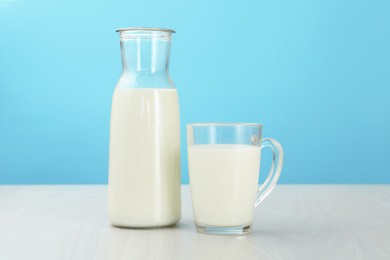 Photo of Fresh milk in cup and jug on white wooden table against light blue background