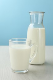 Photo of Fresh milk in cup and jug on white wooden table against light blue background