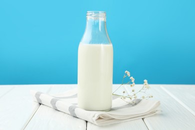 Photo of Fresh milk in glass bottle and flowers on white wooden table against light blue background