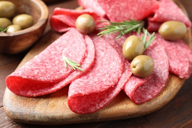 Photo of Slices of delicious sausage with olives and rosemary served on wooden table, closeup