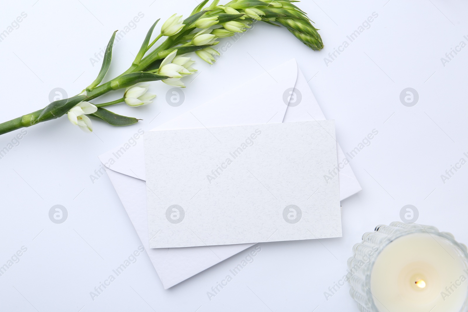 Photo of Blank greeting card, paper envelope, burning candle and flowers on white background, flat lay