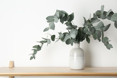 Photo of Beautiful eucalyptus branches in vase on wooden table near white wall indoors, space for text