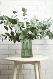 Photo of Green eucalyptus branches in vase on table near white brick wall indoors