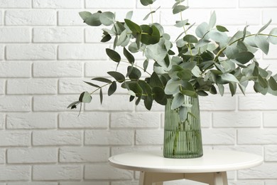 Photo of Green eucalyptus branches in vase on table near white brick wall indoors, space for text