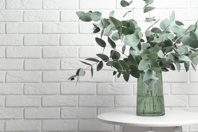 Photo of Green eucalyptus branches in vase on table near white brick wall indoors, space for text
