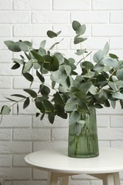 Photo of Green eucalyptus branches in vase on table near white brick wall indoors