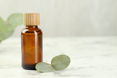 Photo of Bottle of essential oil and eucalyptus leaves on white marble table, closeup. Space for text
