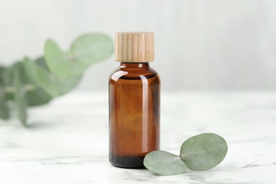 Photo of Bottle of essential oil and eucalyptus leaves on white marble table, closeup