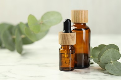 Photo of Bottles of essential oil and eucalyptus leaves on white marble table, closeup. Space for text