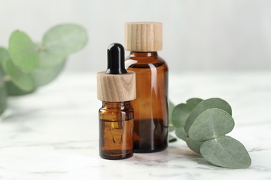 Photo of Bottles of essential oil and eucalyptus leaves on white marble table, closeup