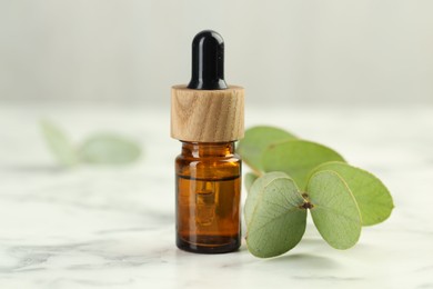 Photo of Bottle of essential oil and eucalyptus leaves on white marble table, closeup