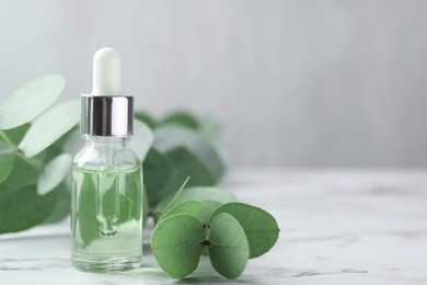 Photo of Bottle of essential oil and eucalyptus leaves on white marble table, closeup. Space for text