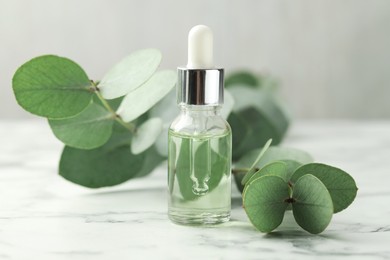 Photo of Bottle of essential oil and eucalyptus leaves on white marble table, closeup