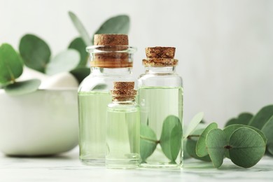 Photo of Bottles of essential oil, eucalyptus leaves, mortar and pestle on white marble table, closeup